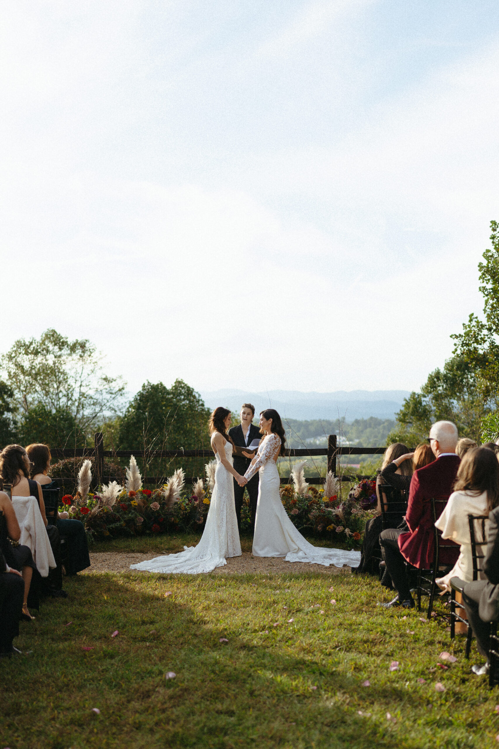Pictured is a wedding photo by Laura Cate Photography, featuring a lesbian couple at their wedding in North Carolina outside on green grass and a blue sky and mountain background.